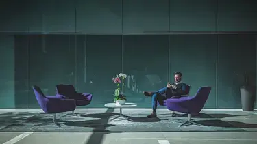 A business leader dressed in a blue suit, sits cross-legged on a couch in a lounge, looking at his mobile device.